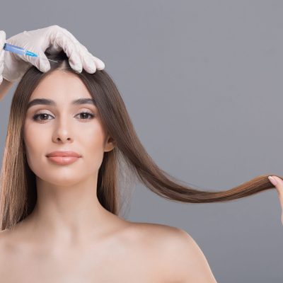 Woman with long brown hair receiving a cosmetic scalp injection: a gloved hand holds a syringe near her hairline.